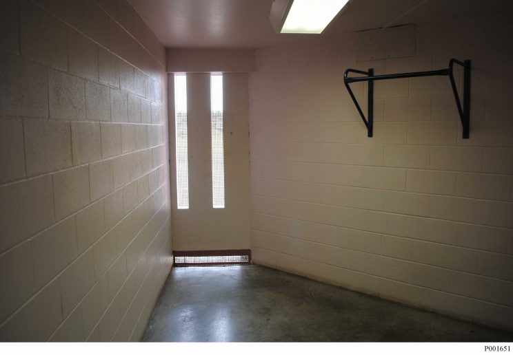 A cinderblock room with walls that narrow toward the far end from approximately 8 feet wide near the photographer to approximately 3 feet wide at the far end. At the end, there are two long narrow windows extending from about one foot off the floor to about 6 inches from the ceiling. The room is empty except for a flourescent light on the ceiling and a chin-up bar on the right wall.