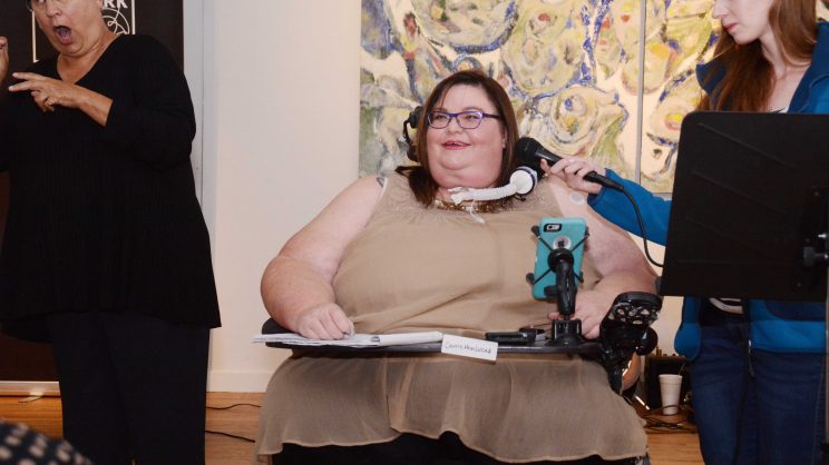 Image: Carrie Ann Lucas, light skinned Latina woman of size with mid-length brown hair, wearing a beige blouse and dark skirt. She is speaking into a mic held for her by a white woman with red hair. To the left, a woman dressed entirely in black interprets into ASL.