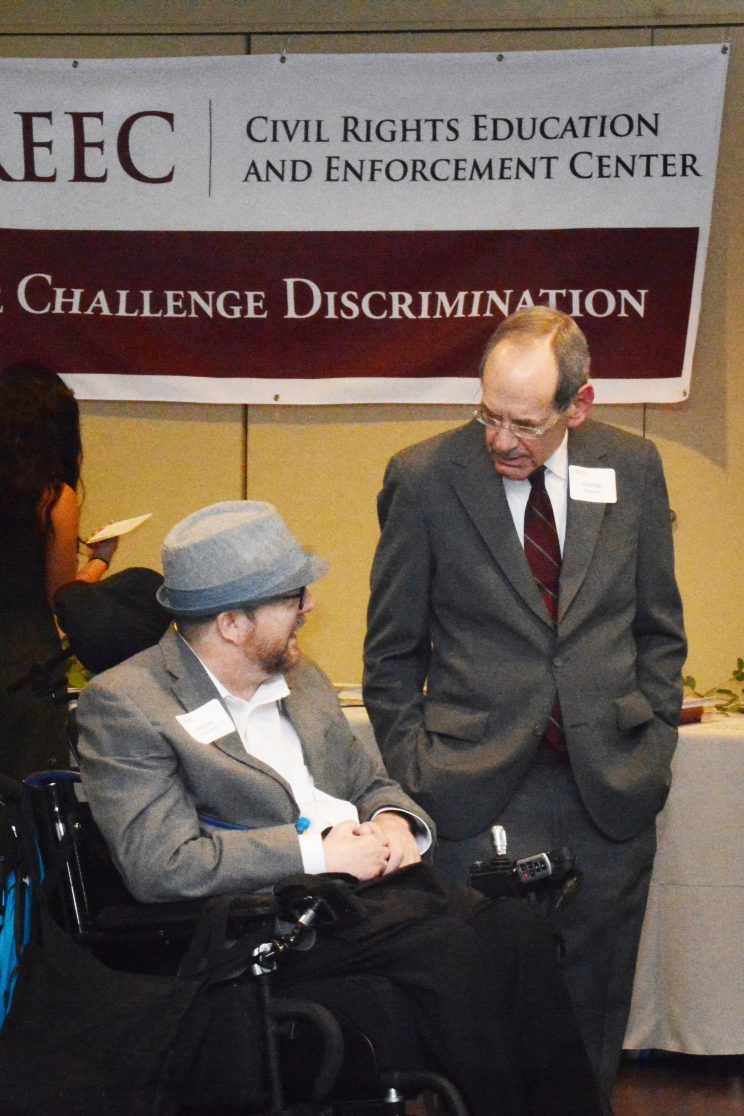 Two men talking. One is standing and one is seated in a wheelchair.