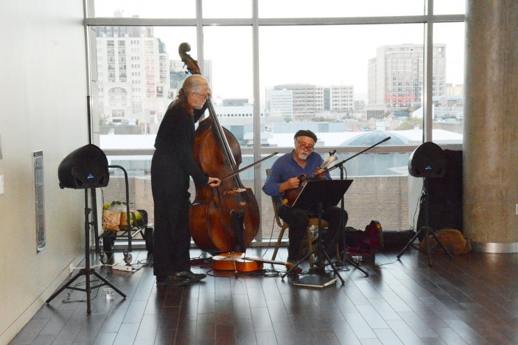 Image of musicians playing standup bass and smaller string instrument in front of large windows showcasing Downtown Denver.