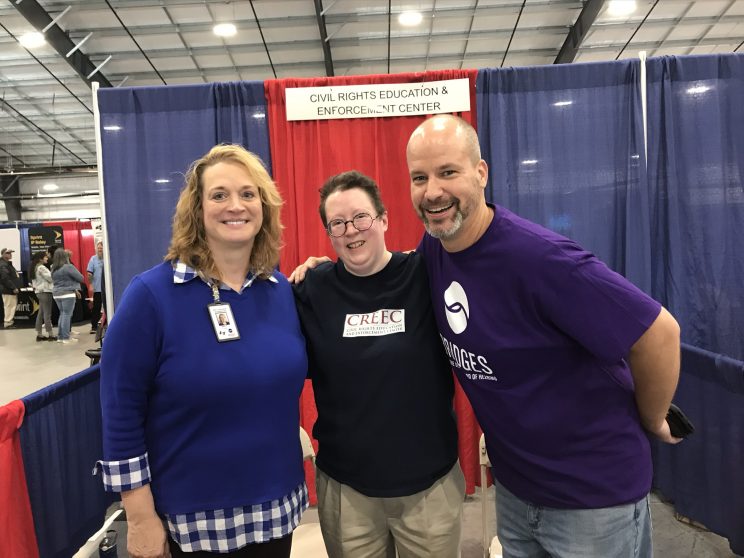 Two white women and one white man. Martie is in the middle wearing a blue shirt with the Disability Law United logo. Our interpreter for the event, Beth Pilkington, is on the left in a blue shirt. On the right in a purple shirt is Mike Helms, VP of Adult Education and Outreach at Bridges in Nashville. Behind the trio is the Disability Law United booth with red and blue curtains and a sign that says Disability Law United.
