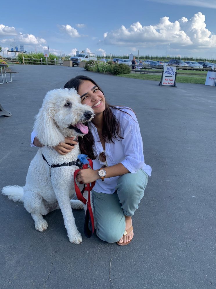 Parima Kadikar is kneeling next to and hugging her dog (a white labradoodle) in a parking lot. Parima is wearing jeans, sandals and a purple oxford shirt. 