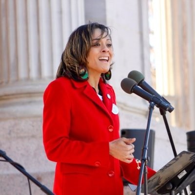 Elisabeth Epps is standing in front of a microphone giving a speech. She is wearing a red blazer.