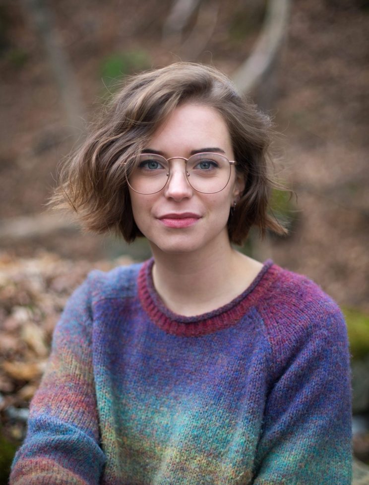 Person with glasses and short brown hair wearing a multicolored sweater outdoors.