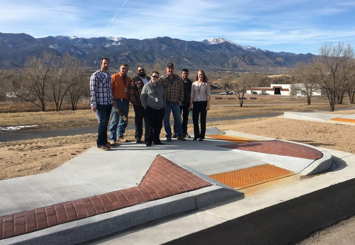 Photograph of seven individuals, including Disability Law United staff member Martie Lafferty and Colorado Springs city staff, standing on pavement in front of mountains. The pavement has a large brick and concrete curb with an on-ramp and textured area. 