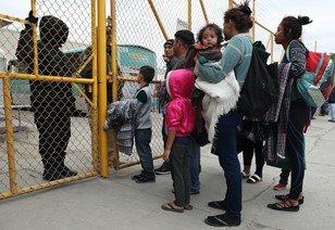A group of about six individuals, including adults and children, standing in front of a yellow gate. There are two figures dressed in dark clothing behind the yellow gate.
