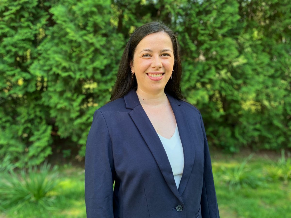 Photograph of Ruby. Ruby is a light skinned person with long dark hair. She is wearing a navy blazer with a pale blue shirt underneath. She is smiling and standing in front of a wall of green trees.