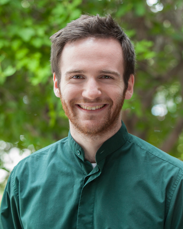 Photo of Sam, a person with pale skin, short brown hair, and a short brown beard. They are wearing a dark green button-up shirt and smiling.