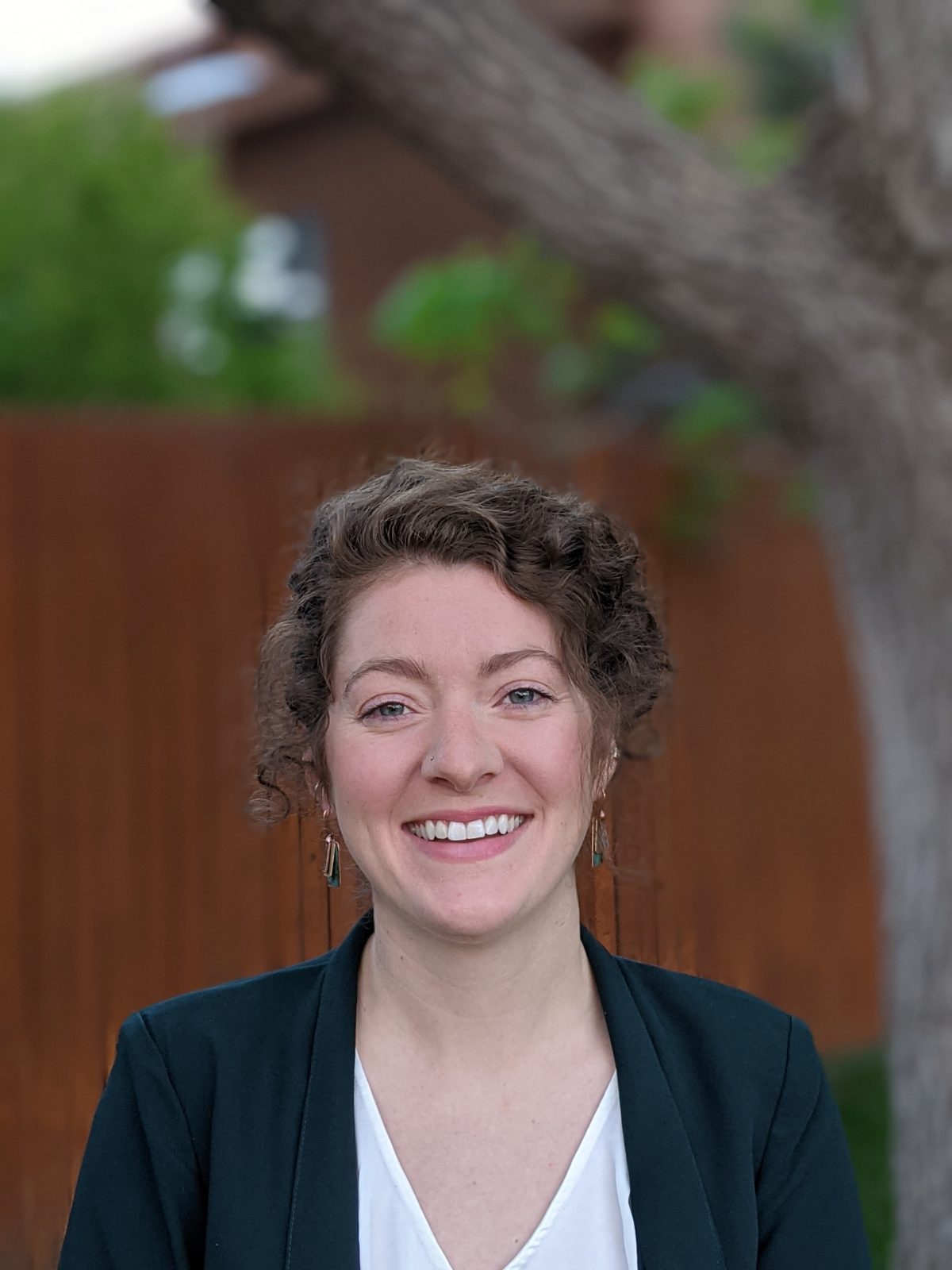Picture of Molly O'Hara. Molly is a person with pale skin and brown curly hair which is short. She is wearing earrings, a dark blazer, and a white shirt. She is smiling. The background is blurry but shows a tree and the side of a building.