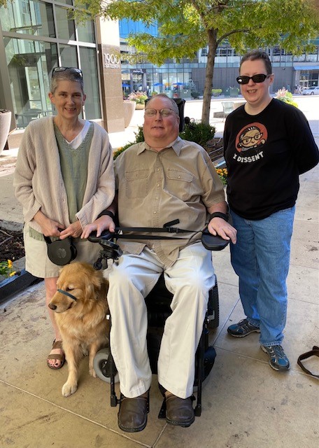 Amy, Tim, Martie, and Holly (dog) smiling together. Amy is a white woman with short gray hair. Tim is a white man with short hair and glasses in a wheelchair. Martie is a white woman with short dark hair and sunglasses. Holly is a golden retriever.
