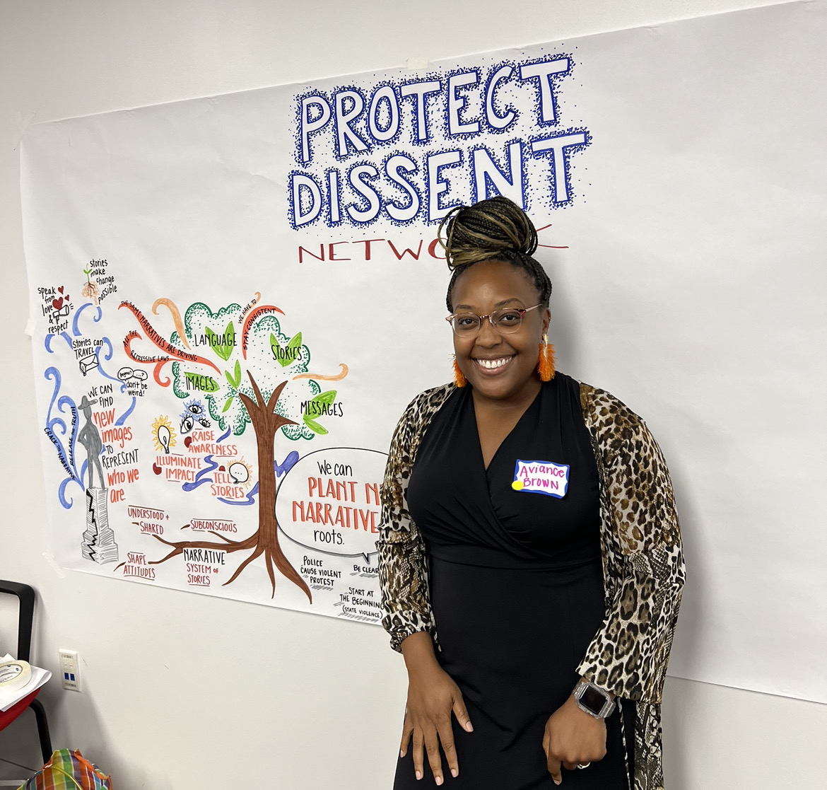Photo of Aviance standing in front of a wall with a large piece of white paper on it. Text at the top of the paper reads: ''Protect Dissent Network.'' There is a drawing of a tree with words surrounding it including: "Images," "Language," "Stories," and "Messages."