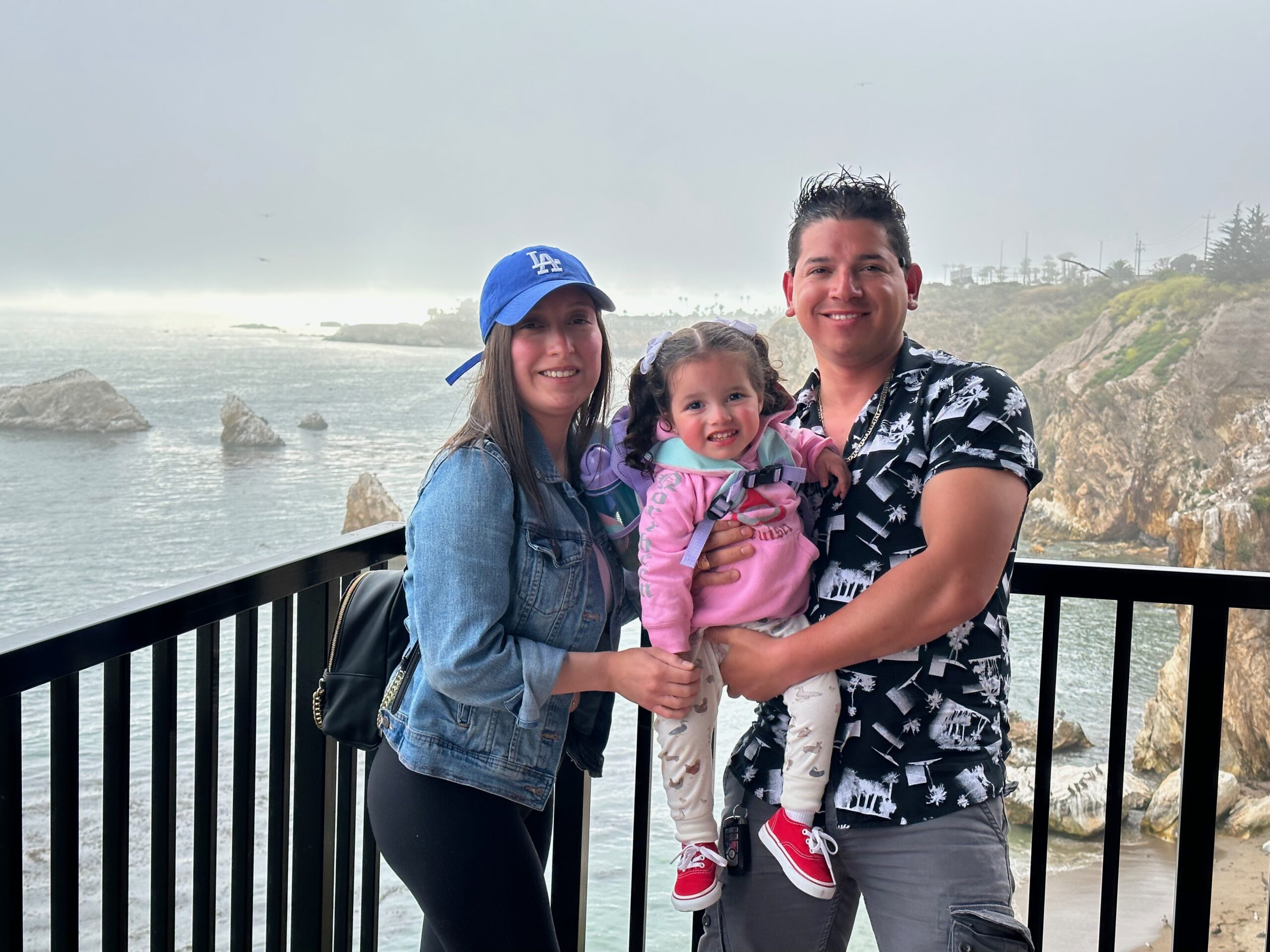 Image of Ulises (on the right) with his wife and daughter. They are stahdning along a metal deck railing overlookcing the ocean. They are all smiling at the camera, looking happy.